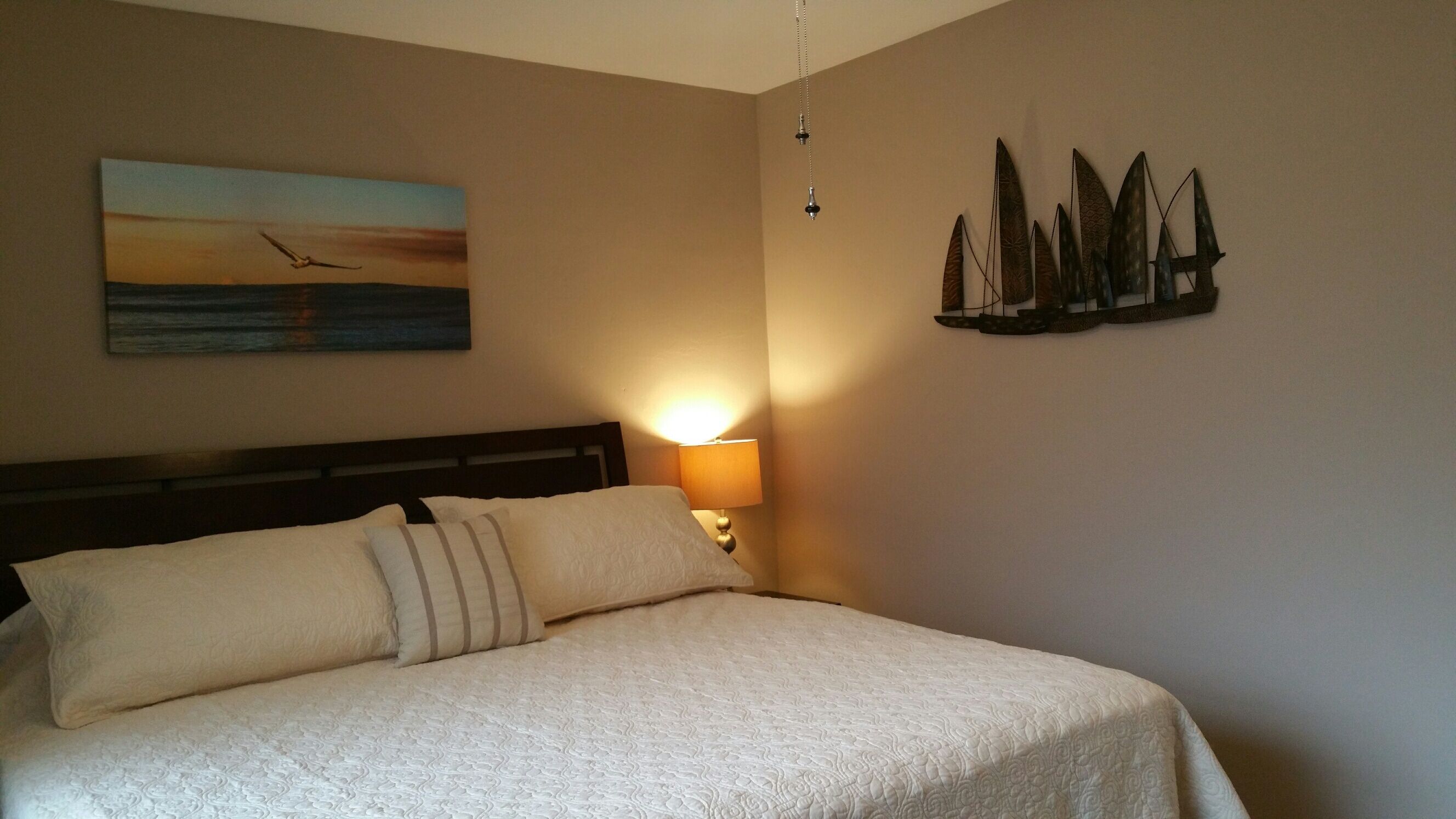 A neatly made bed with a headboard, a bedside lamp, a painting of a sunset over water, and a wall-mounted decorative piece.