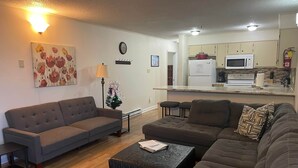 A living room with a sofa, ottoman, and a small table. There is a kitchen area with cabinets and appliances. - Winterplace Condo D303 (Ghent)