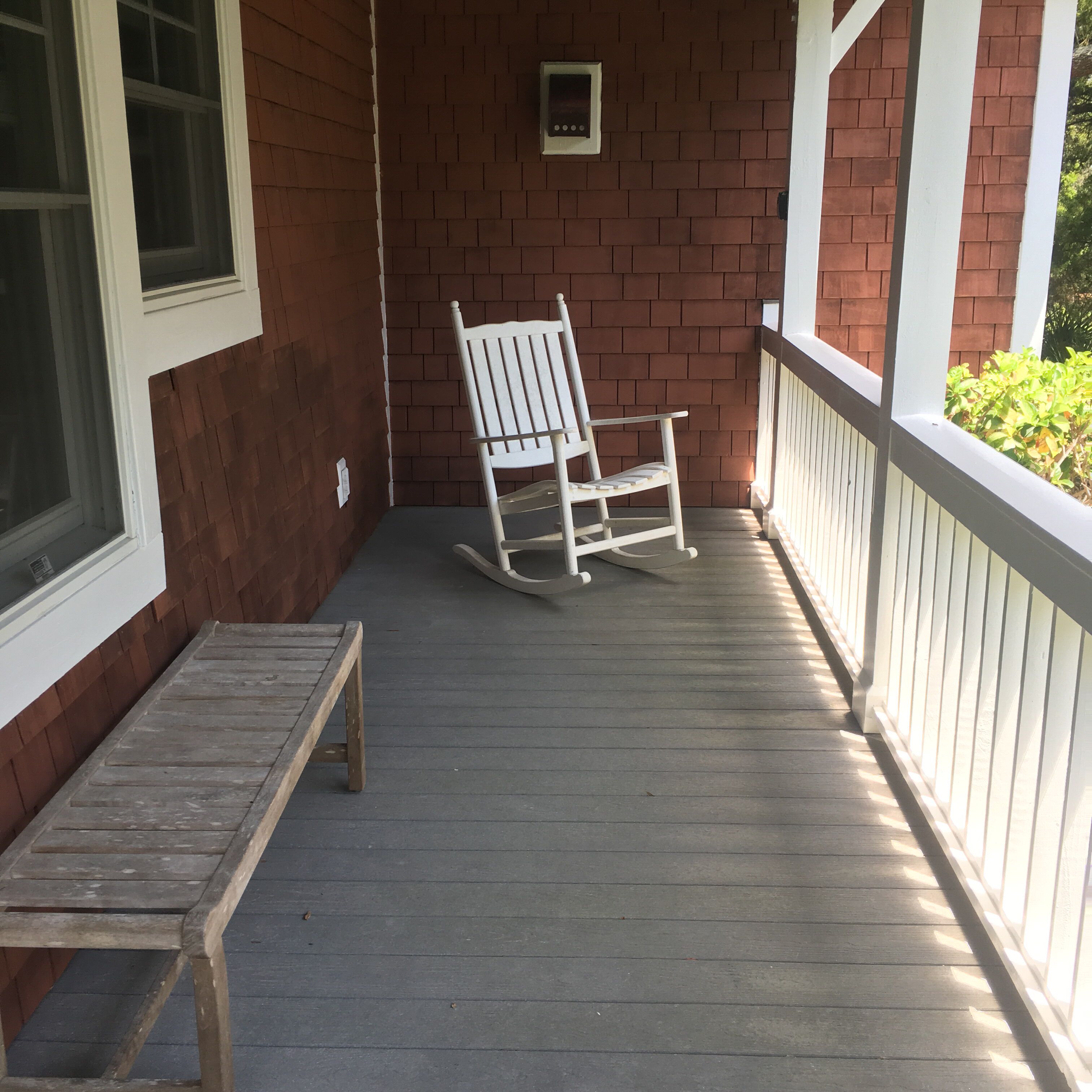 A wooden rocking chair on a porch with a brick wall and a window.
