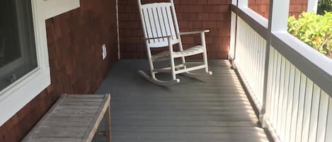 A wooden rocking chair on a porch with a brick wall and a window.