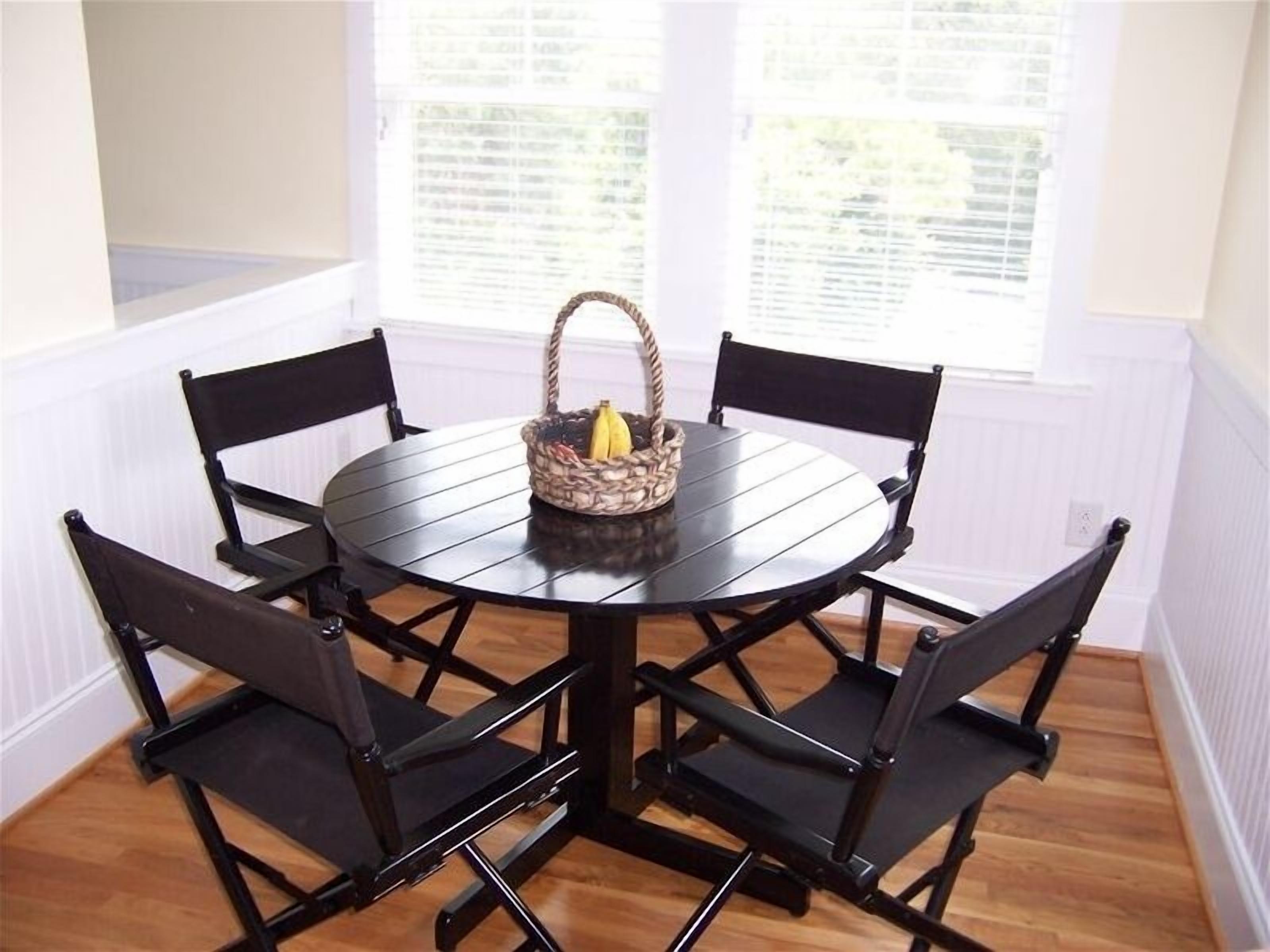 Una mesa de comedor redonda con cuatro sillas negras, una cesta tejida con plátanos y una ventana con persianas de fondo.