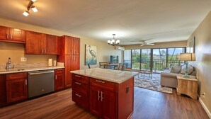 A spacious kitchen with wooden cabinets, a dishwasher, and a large island. There is a dining area with a table and chairs, and a balcony with a view. - Steps to beach! Beautiful renovated 1 bedroom (Kihei)