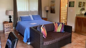 Une chambre à coucher comprenant un lit, un canapé en osier, une table de chevet, une lampe et une salle de bain visible à travers une porte en verre.