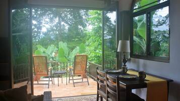 A room with a view of lush greenery through large windows, two wooden chairs, a small table with a lamp, and a patterned rug.