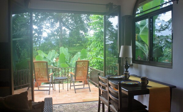 A room with a view of lush greenery through large windows, two wooden chairs, a small table with a lamp, and a patterned rug.
