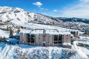 A multi-story hotel with a snow-covered roof, situated in a mountainous area with a ski resort in the background.