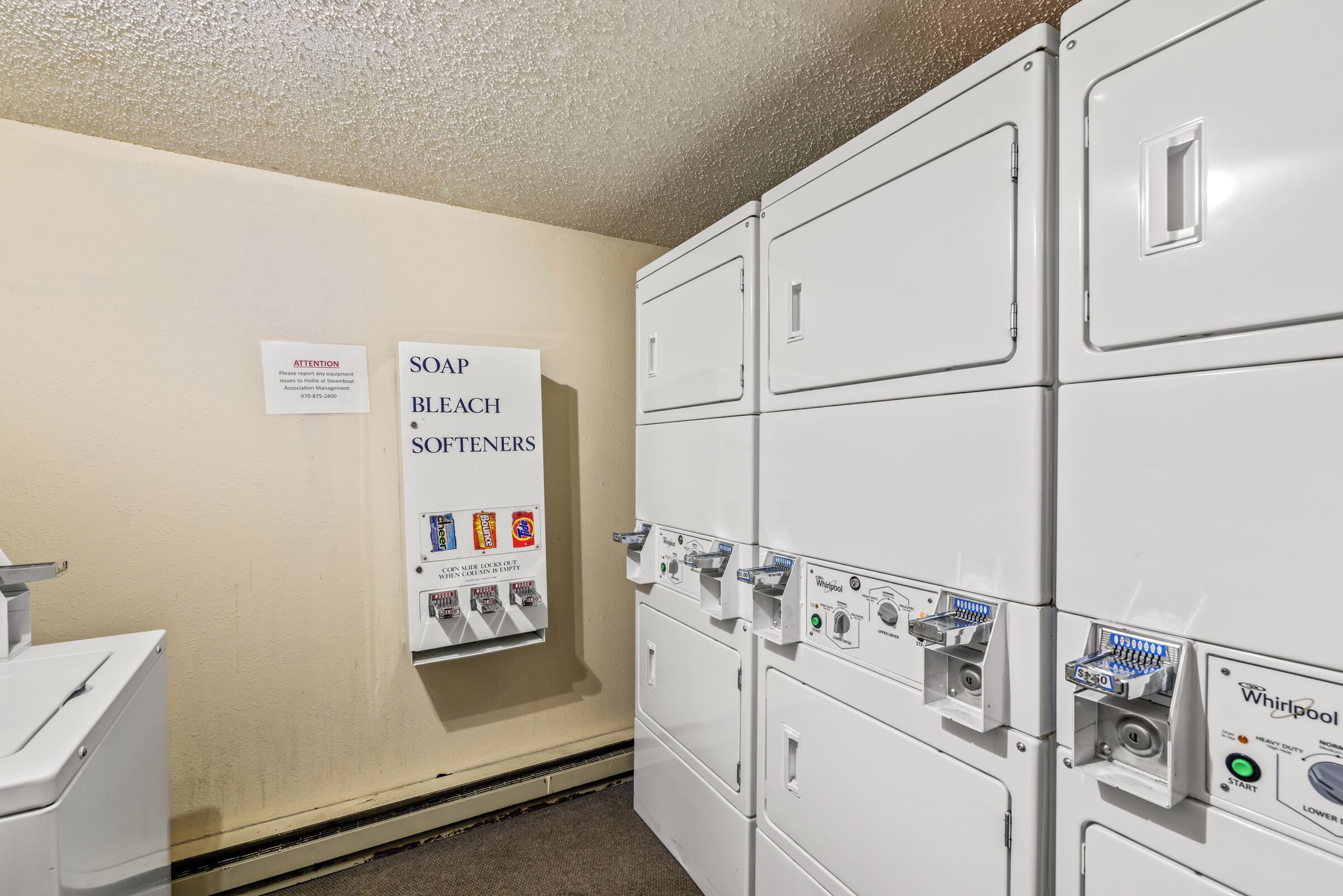 Un espace dédié à la buanderie, équipé de machines à laver et de sèche-linge superposés, d’un distributeur de lessive, d’adoucissant et d’eau de Javel, ainsi que d’un panneau d’avertissement.