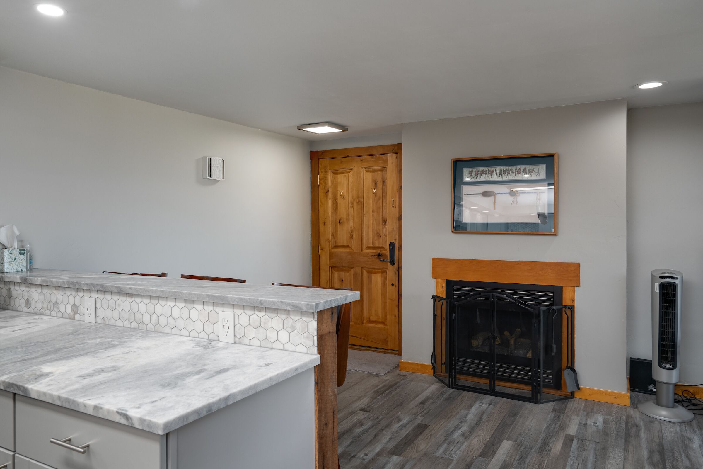 A kitchen with a marble countertop, wooden cabinets, a hexagonal tile backsplash, a wooden door, a fireplace, and a standing fan.