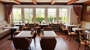 A restaurant with multiple dining tables and chairs, large windows with curtains, and a view of greenery outside.
