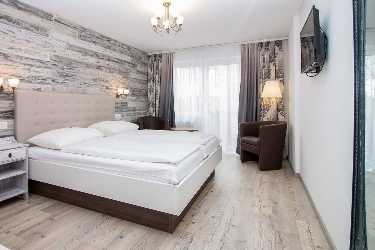 A modern hotel room with a wooden wall, a large bed, bedside tables, a chair, and a TV mounted on the wall.