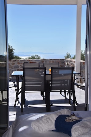 A balcony with a table and chairs, a bed with a white and blue blanket, and a view of the sea and mountains.
