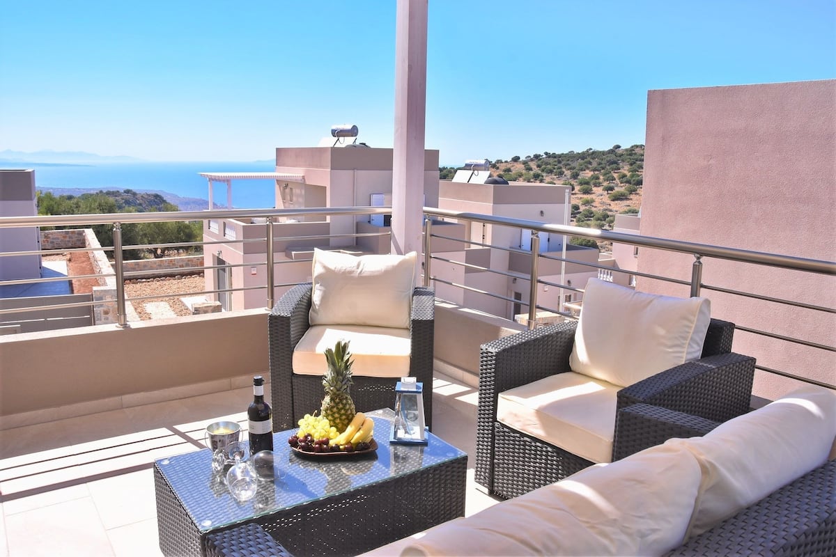 Une terrasse sur le toit, meublée de mobilier en osier, avec une table en verre sur laquelle est posé un panier de fruits, et d’où l’on a une vue dégagée sur la mer et les collines à l’arrière-plan.