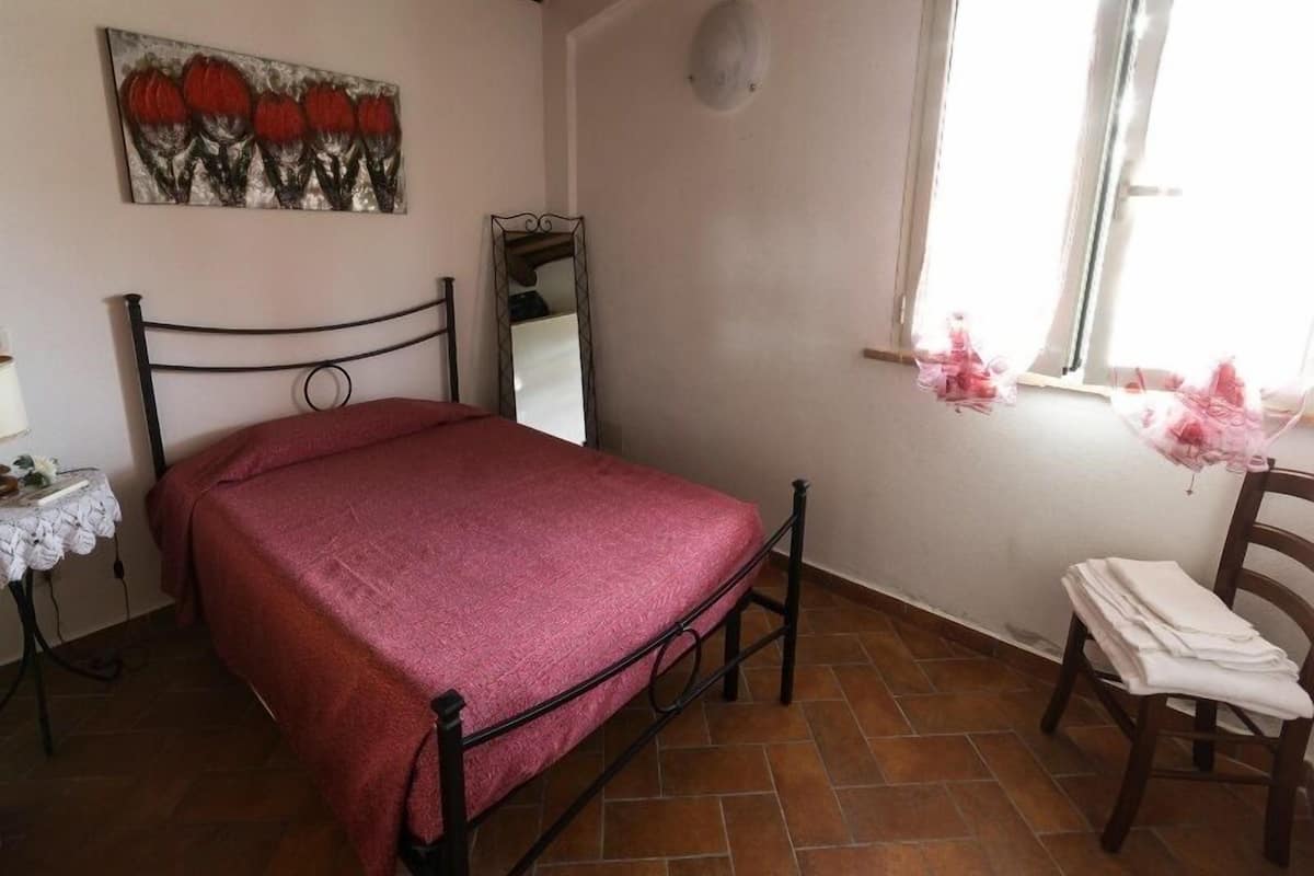 A bedroom with a bed, a bedside table, a mirror, a window with curtains, and a chair with folded towels.