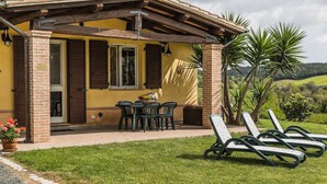 Ein gelbes Gebäude mit einer Ziegelstütze, einer überdachten Terrasse, einem Tisch mit Stühlen und einer Reihe von Liegestühlen.