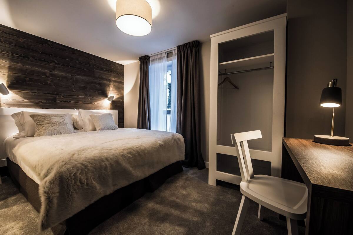 Une chambre moderne avec un grand lit, un bureau et une chaise. On y trouve également une armoire et une fenêtre avec des rideaux.