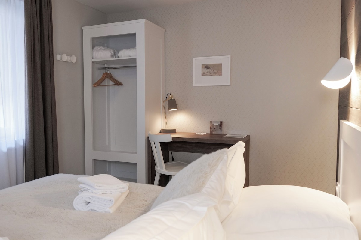 A neatly made bed with a white comforter and folded towels, a wooden nightstand with a lamp, a white wardrobe, and a framed picture on the wall.