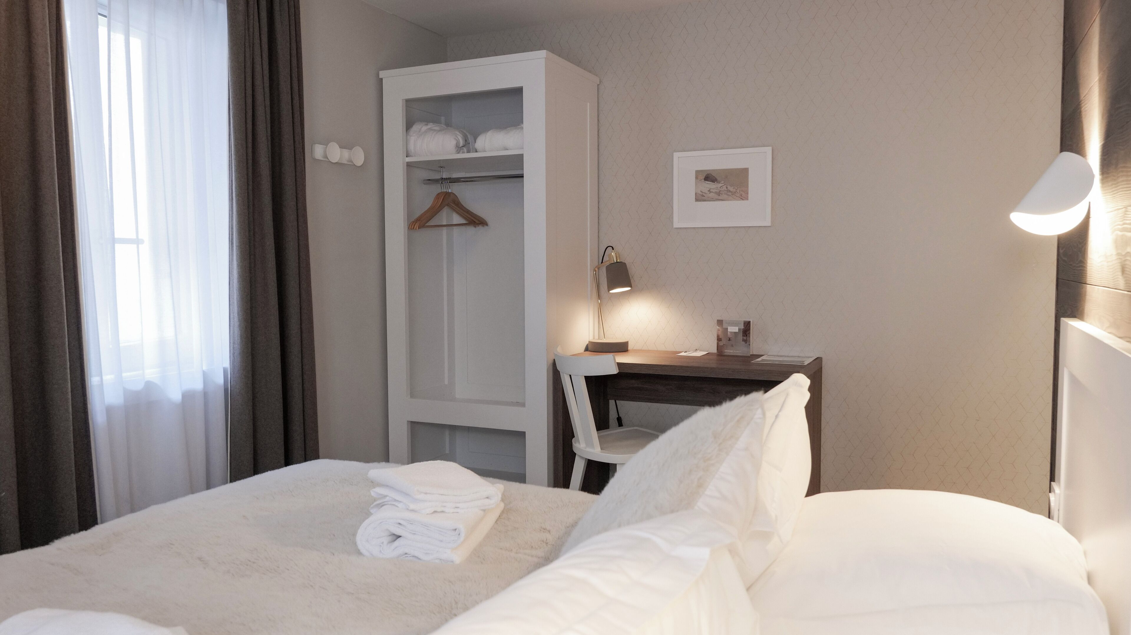 Un lit bien fait, avec du linge de lit blanc, une table de chevet avec une lampe, une armoire et un tableau encadré au mur.