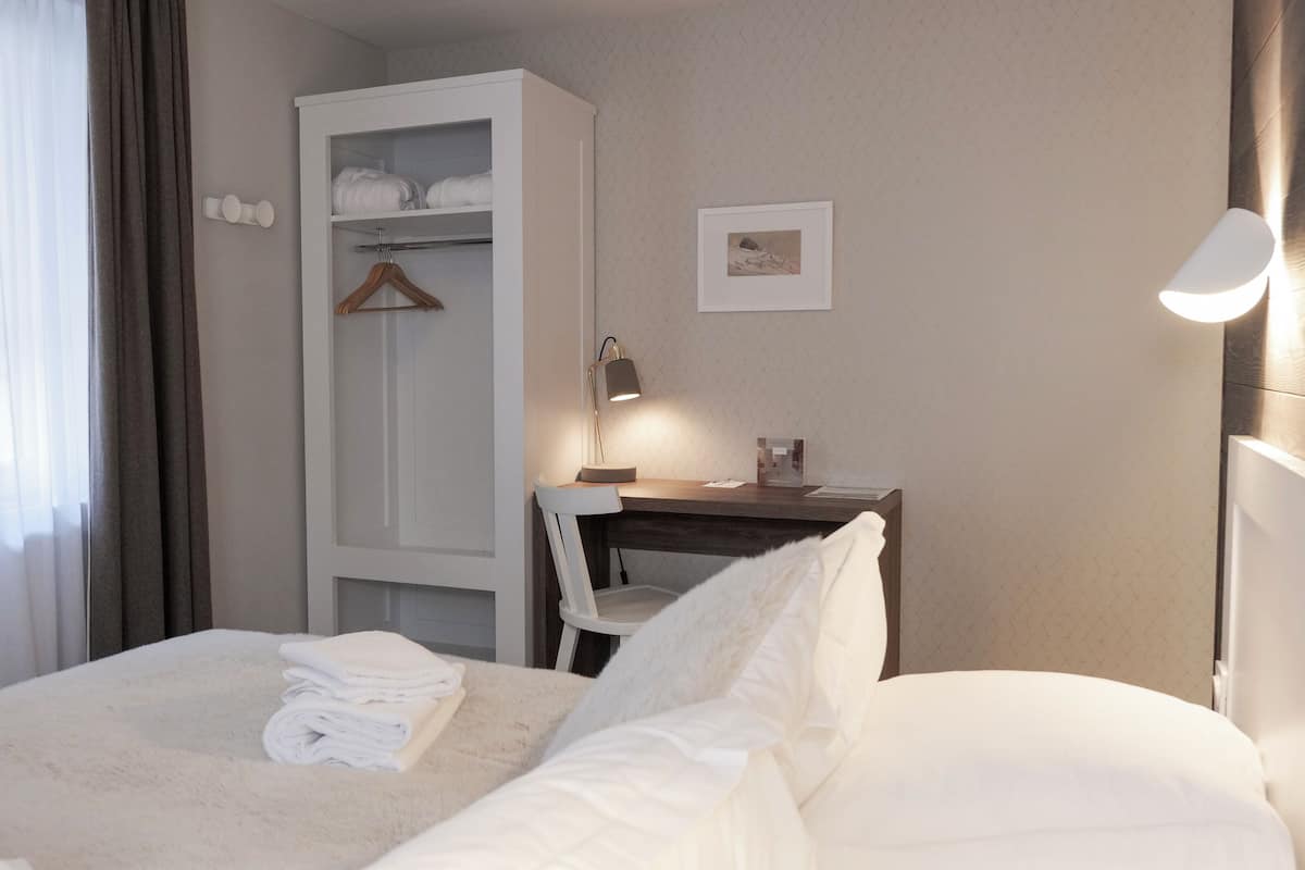 Un lit bien fait, avec une couette blanche et des serviettes pliées, une table de chevet en bois avec une lampe, une armoire blanche et un tableau encadré au mur.