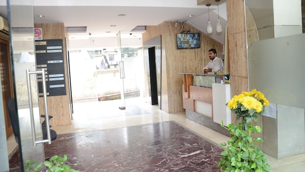 A reception area with a receptionist, a reception desk, a TV mounted on the wall, and a potted plant with yellow flowers. - Hotel Sai Leela Grand (Mumbai)