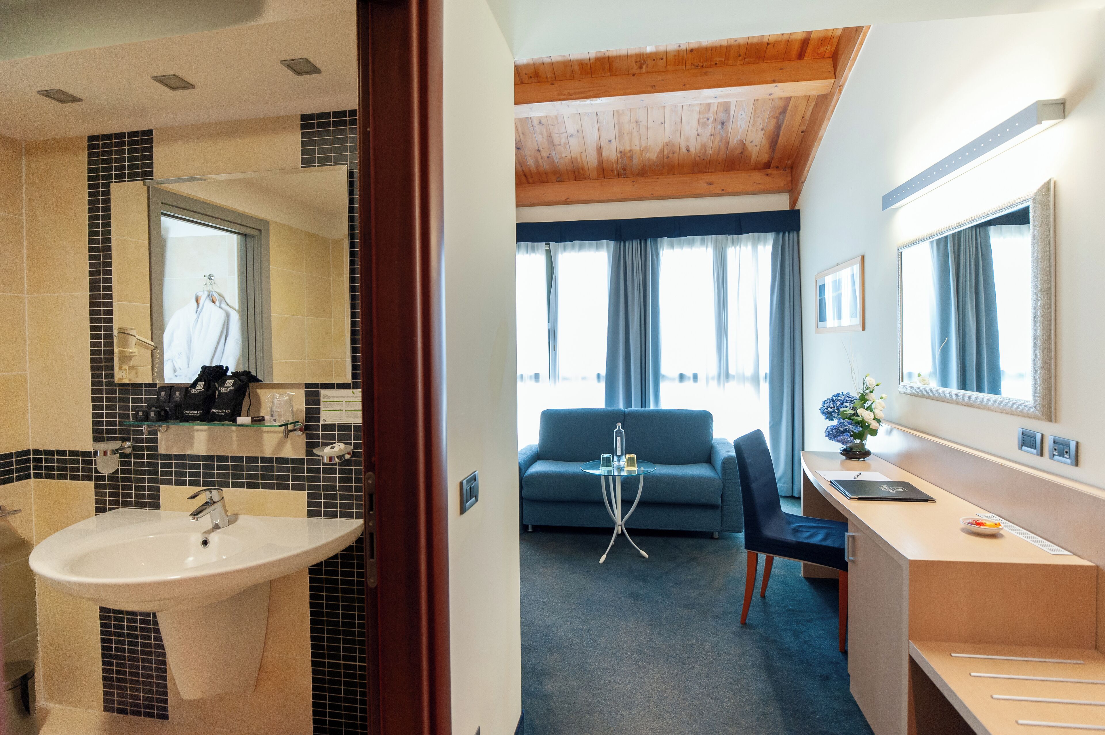 A hotel room with a bathroom, a sofa, a desk, and a large window.
