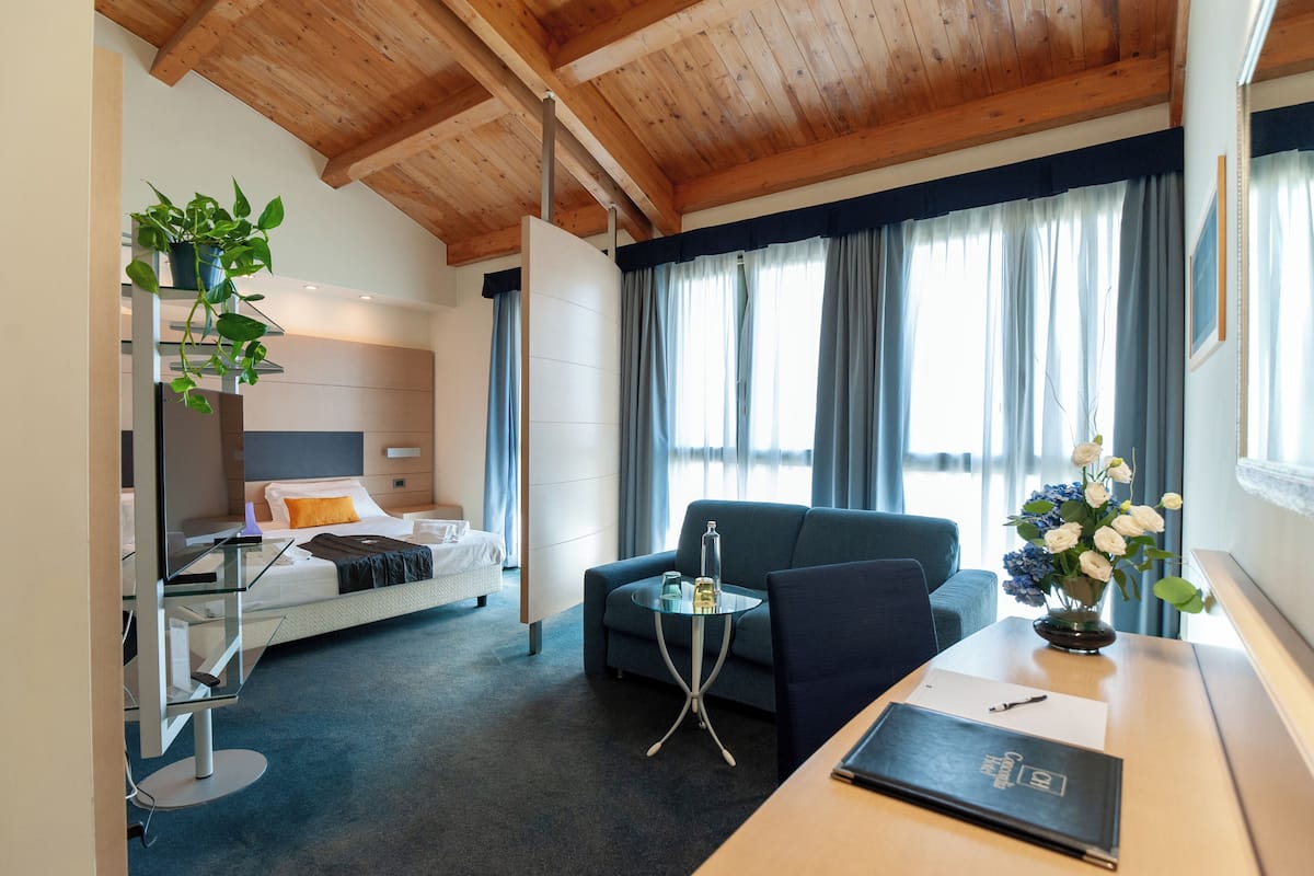 A modern hotel room with a bed, a desk, a chair, a TV, and a plant.