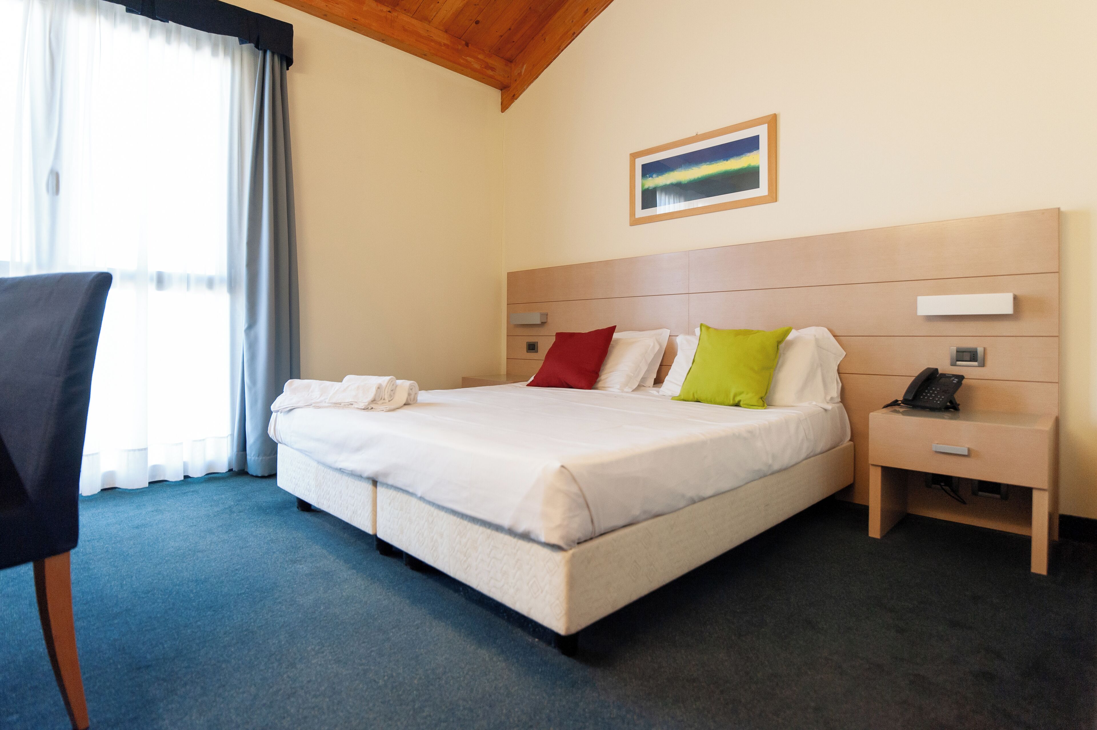 A hotel room with a bed, a chair, a bedside table with a phone, and a painting on the wall.