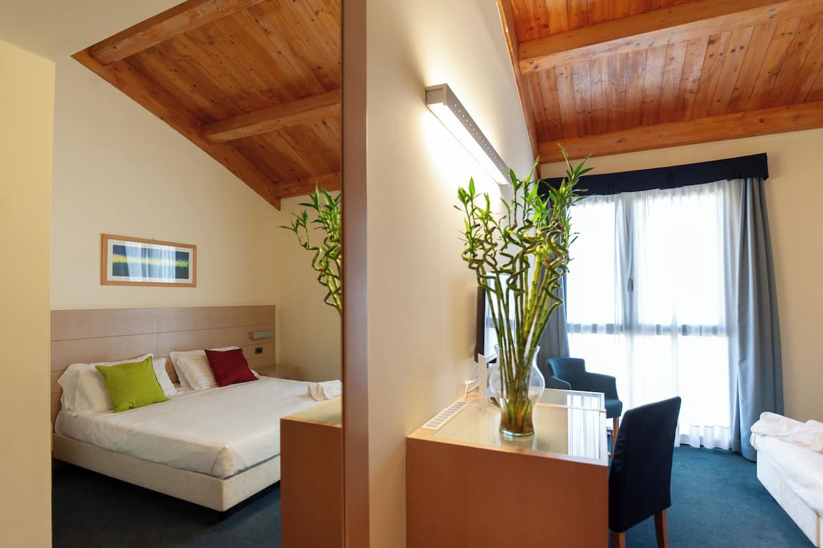 A hotel room with a bed, a desk, a chair, a vase with plants, and a window with curtains.