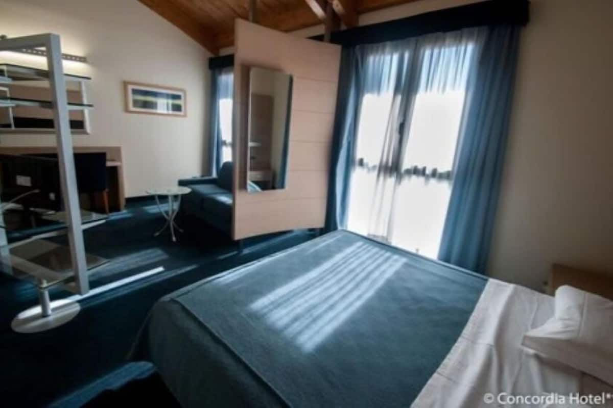 A hotel room with a bed, a desk, a chair, a small table, a wardrobe, and a window with curtains.