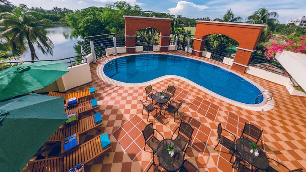 Piscina con suelo a cuadros, rodeada de sillones de baño y sombrillas, con vista a un lago y palmeras.