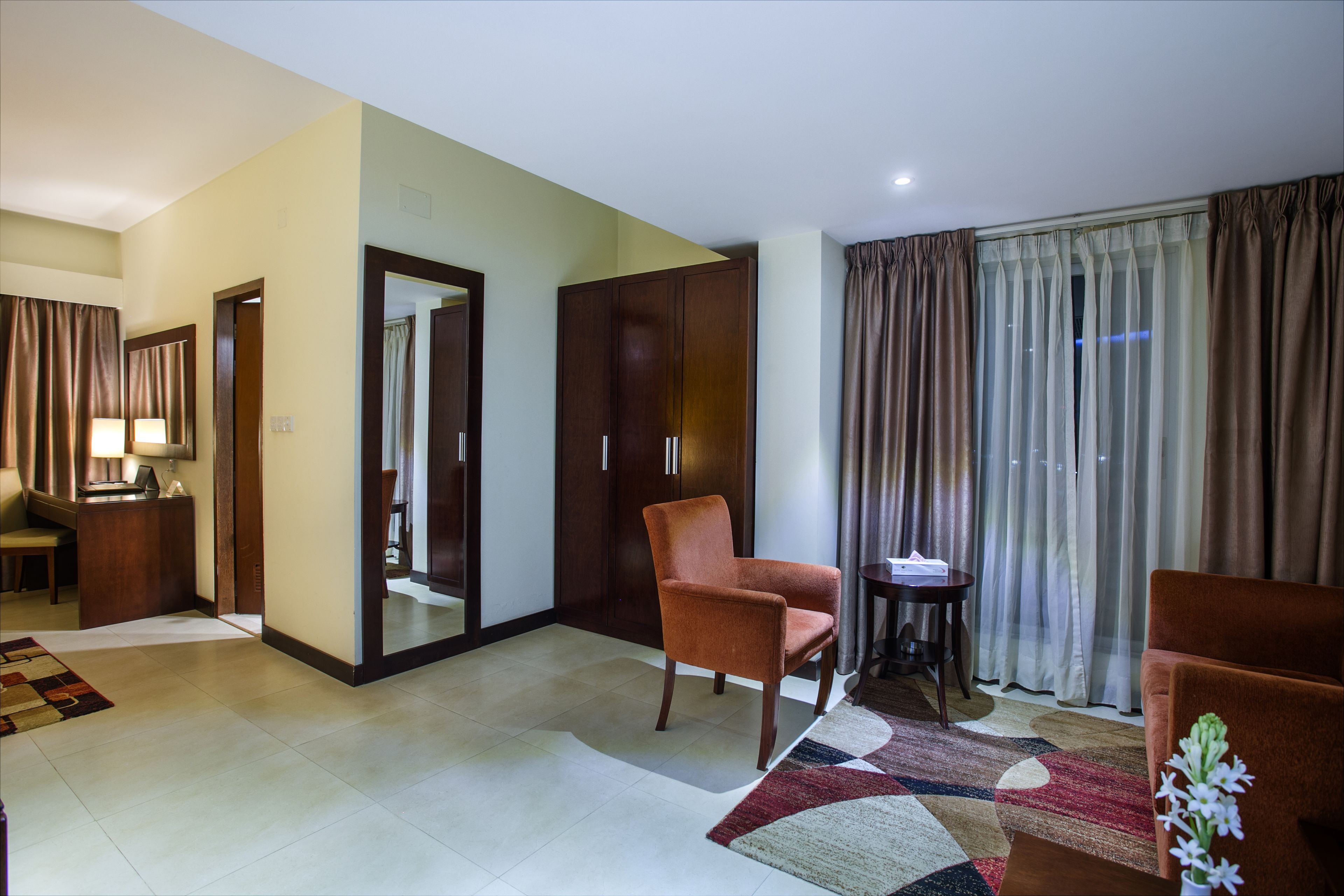 Quarto de hotel com um espelho grande, guarda-roupa de madeira, poltrona marrom, mesa redonda pequena e uma janela com cortinas.