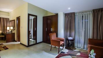 Quarto de hotel com um espelho grande, guarda-roupa de madeira, poltrona marrom, mesa redonda pequena e uma janela com cortinas.