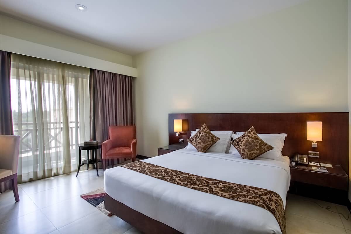 Habitación de hotel con una cama grande, dos lámparas de noche, una mesita de noche, una silla, una pequeña mesa y vistas al exterior a través de cortinas translúcidas.