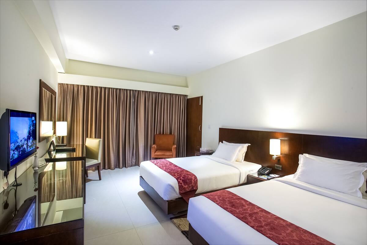 Habitación de hotel con dos camas, televisión, escritorio con computadora, una silla y un amplio ventanal con cortinas.
