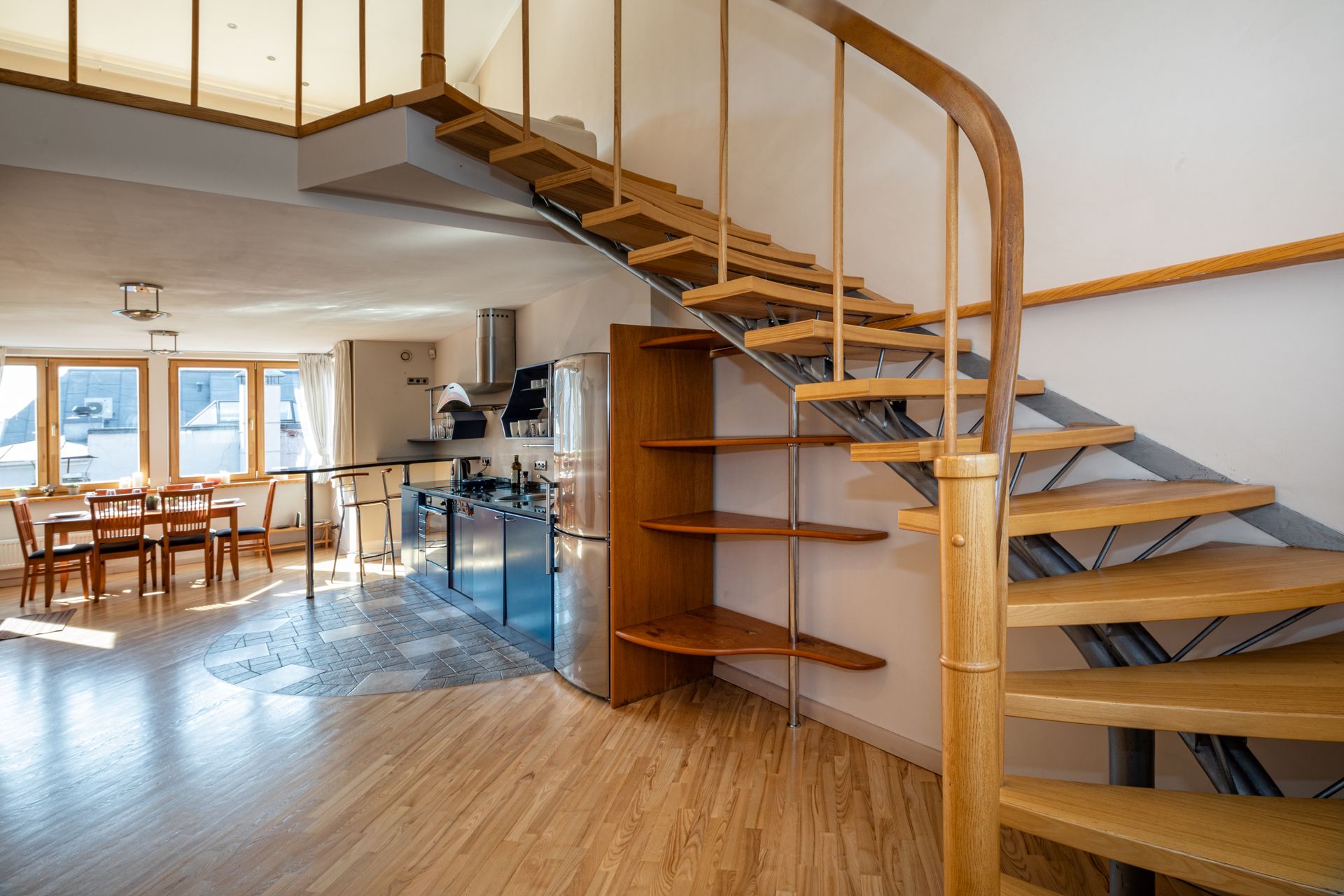 Une cuisine moderne dotée d’un îlot central, d’appareils électroménagers en acier inoxydable et d’un escalier en bois aux lignes courbes.