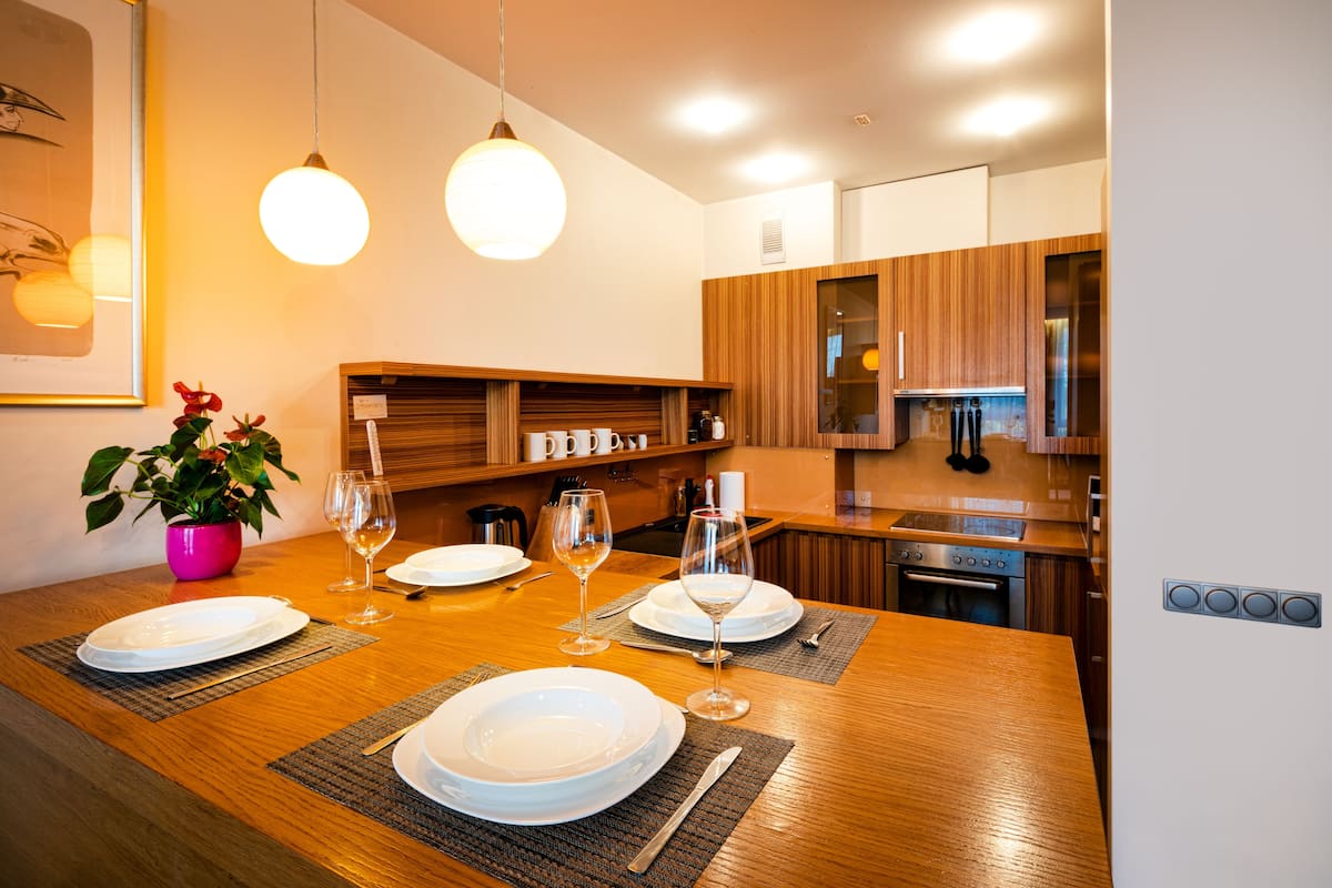 A modern kitchen with wooden cabinets, a dining table set with plates and wine glasses, and a potted plant.