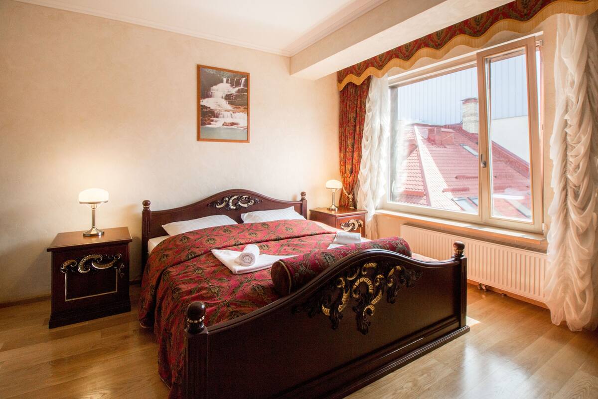 A bedroom with a large bed, two bedside tables, a window with curtains, a radiator, and a painting on the wall.
