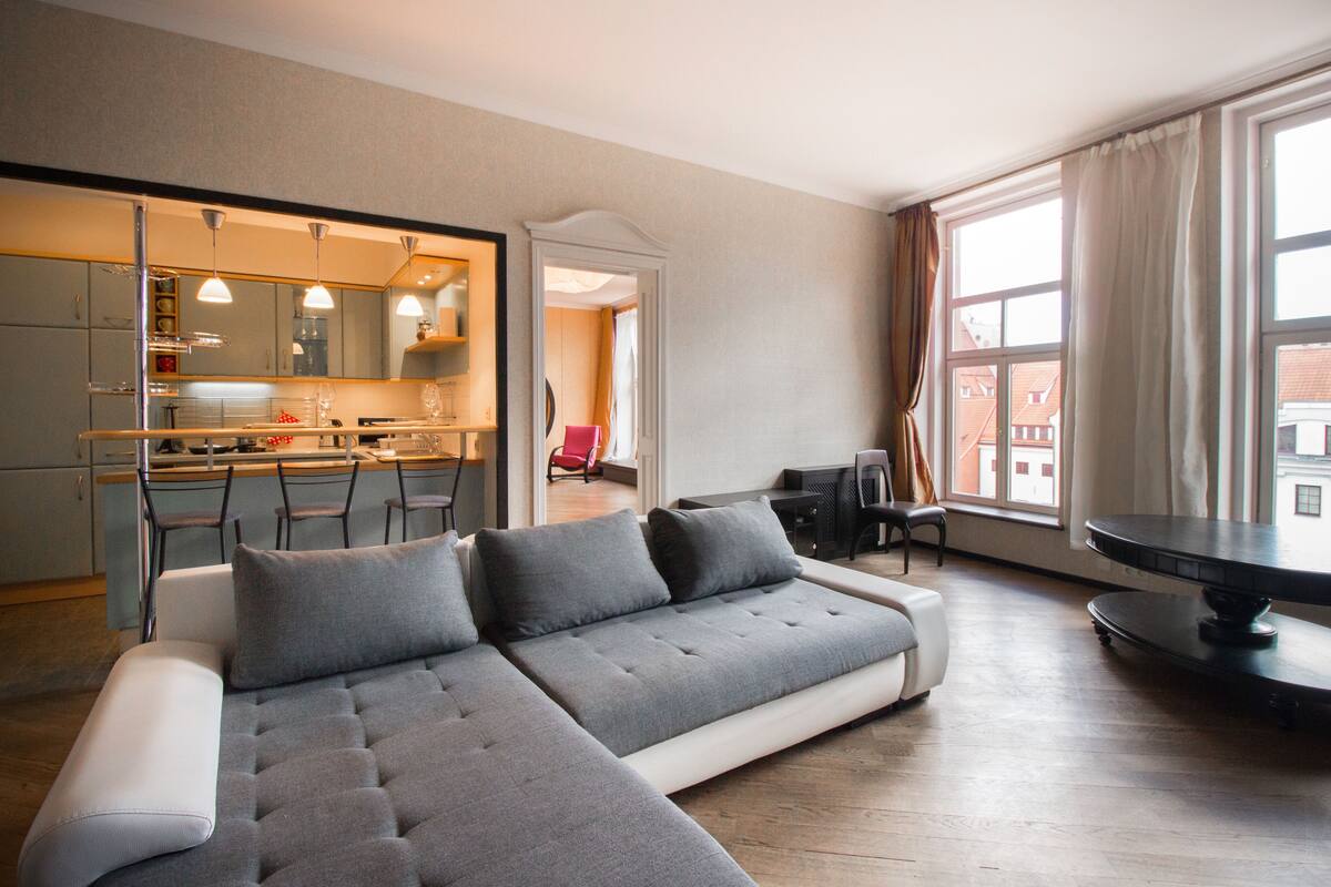 A modern living area with a grey sectional sofa, a dining table, and a kitchen in the background.