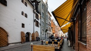A narrow street with outdoor seating under a striped awning, cobblestone pavement, and historic buildings with wooden shutters. - Old Riga Apartments (Riga)