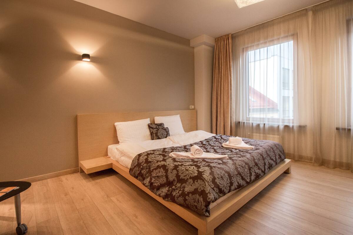 A modern hotel room with a large bed, a bedside table, and a window with sheer curtains.