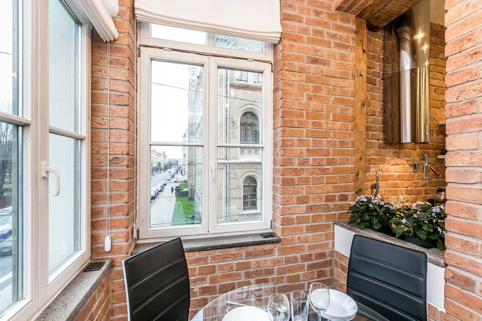 Ein Eckzimmer mit Backsteinwand, einem Fenster mit Blick auf eine Straße, einem kleinen Waschbecken und einem Tisch mit Stühlen.