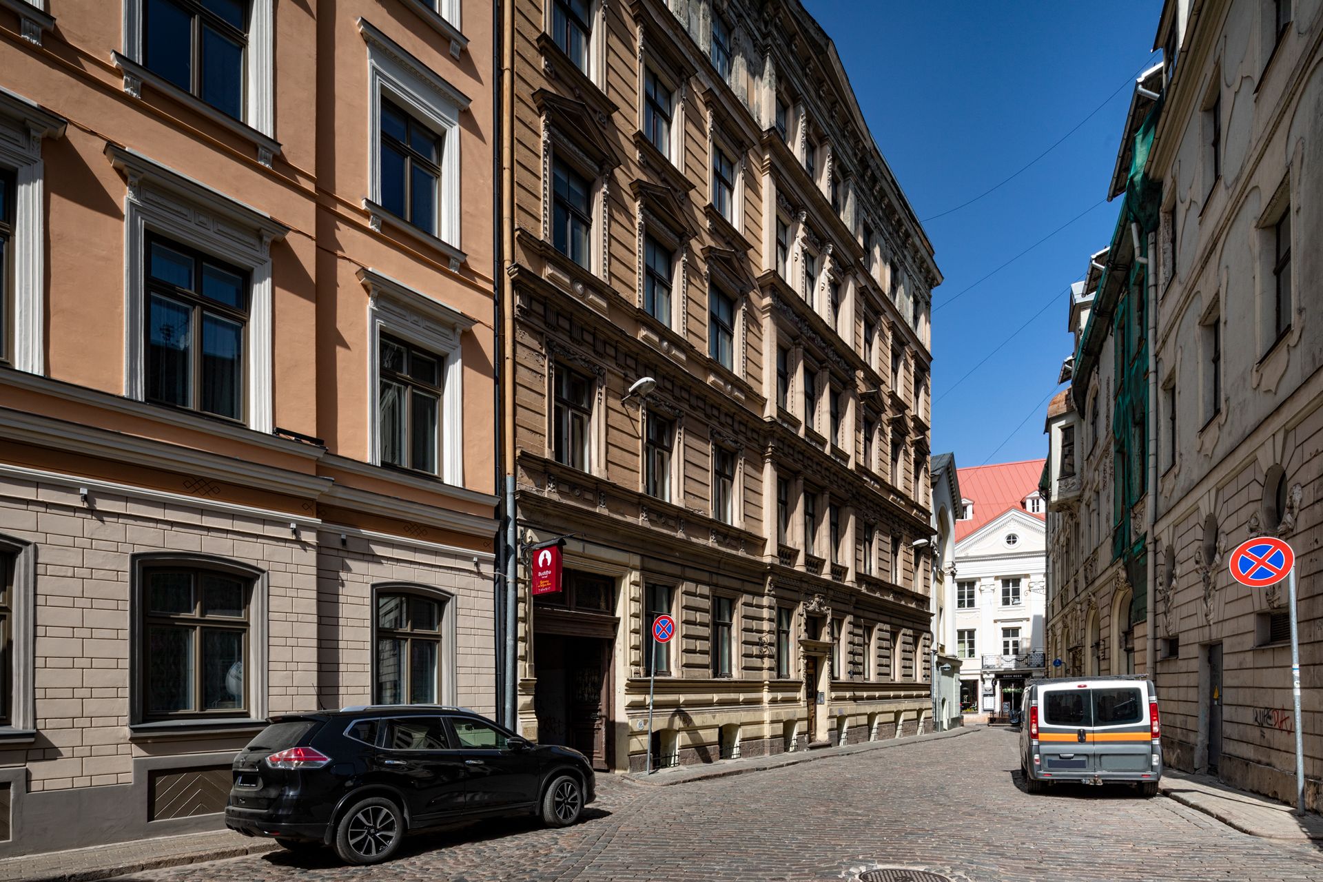 En kullerstensgata kantad av historiska byggnader, en svart bil parkerad vid sidan av vägen och en buss i fjärran.