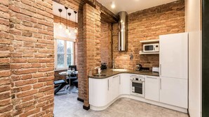 A kitchen with exposed brick walls, white cabinetry, a stainless steel range hood, and a window providing natural light. - Old Riga Apartments (Riga)