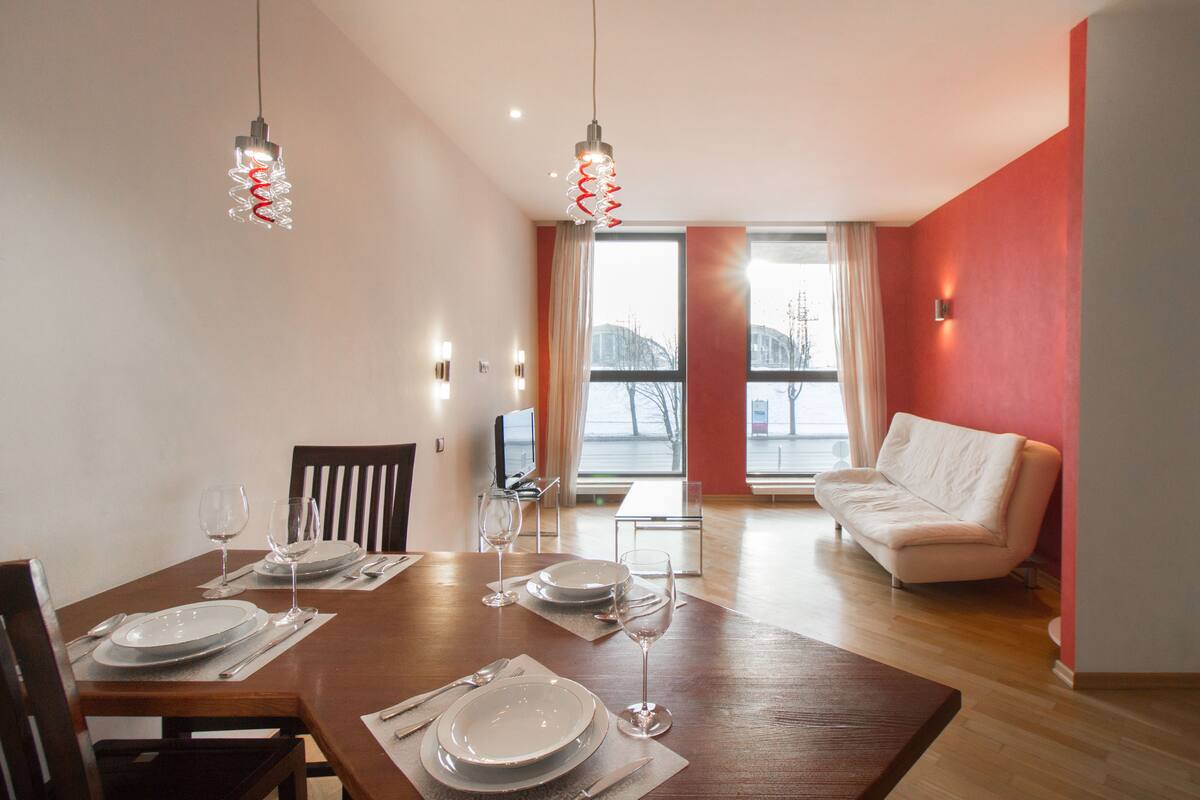 A dining area with a wooden table set for four, two hanging pendant lights, a sofa, and a TV in the background.