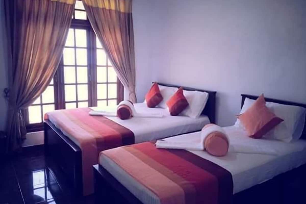 A hotel room with two beds, each with a white mattress and red and orange pillows. The room has a window with curtains and a view of the outside.