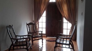 A room with three wooden chairs, a woven rug, and a large window with curtains. - The European Gate (Anuradhapura)