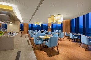 A modern dining area with wooden tables and blue chairs, large windows offering a city view, and contemporary pendant lighting. - Somerset Baitang Suzhou (Suzhou)