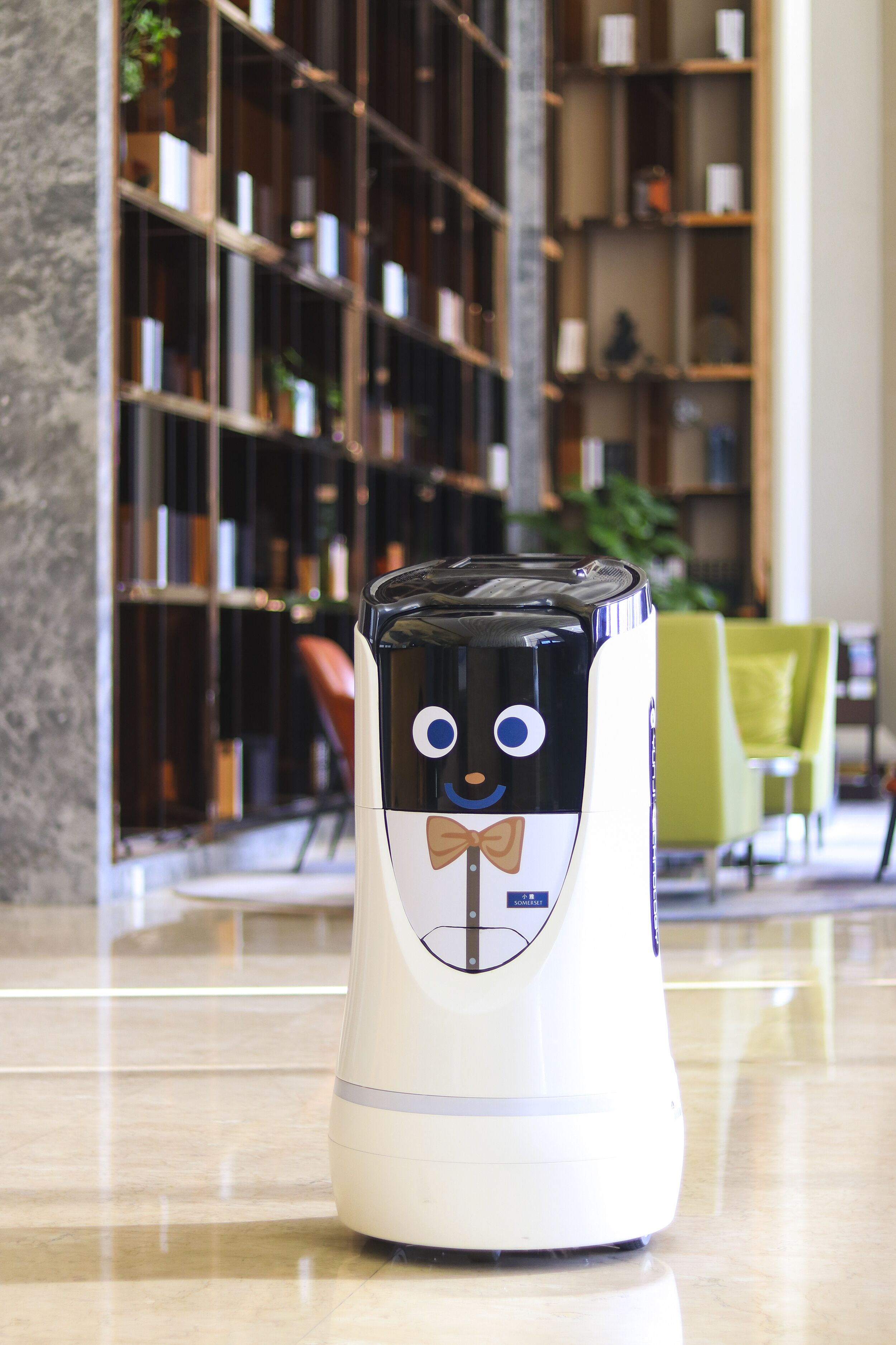A robot with a bow tie standing on a polished floor in a room with a bookshelf and a green chair in the background.