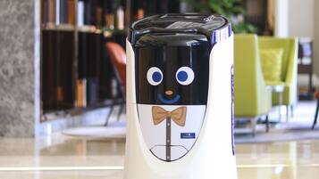 A robot with a bow tie standing on a polished floor in a room with a bookshelf and a green chair in the background.