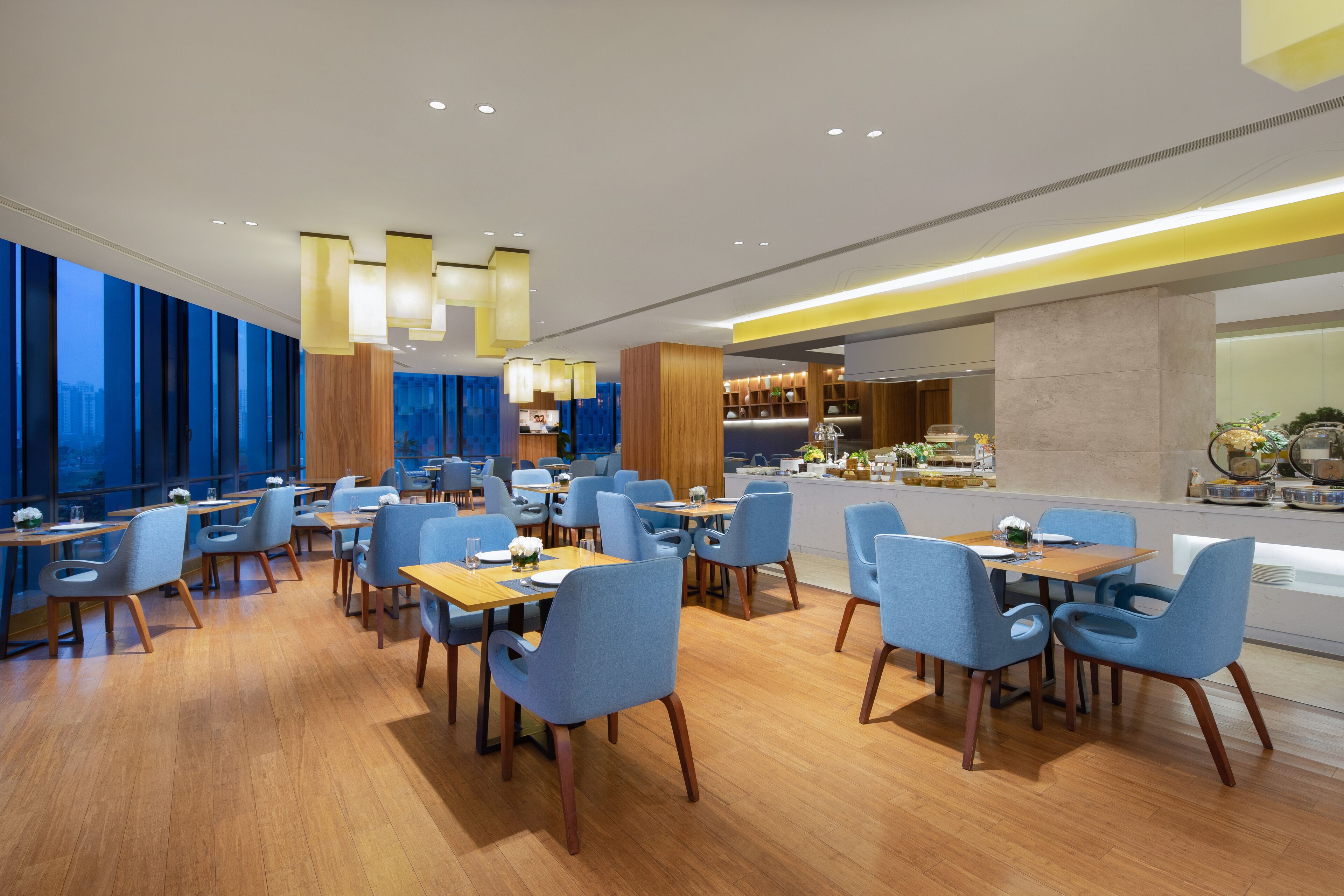 Un restaurante moderno con mesas de madera y sillas azules, una zona de bar y grandes ventanales con vistas a la ciudad.