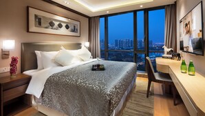A hotel room with a large bed, a desk, a chair, a TV, and a city view. - Somerset Baitang Suzhou (Suzhou)